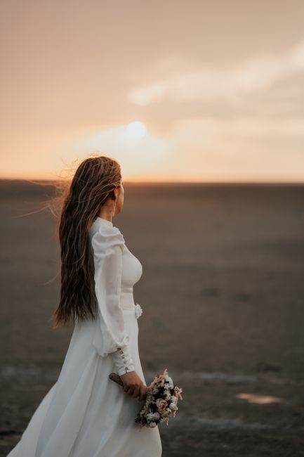 A woman dressed in a white wedding gown stands on a beach during sunset, holdng a bouquet of mixed flowers with pink, cream, and greenery accents. The bouquet features fresh roses, peonies, and small 