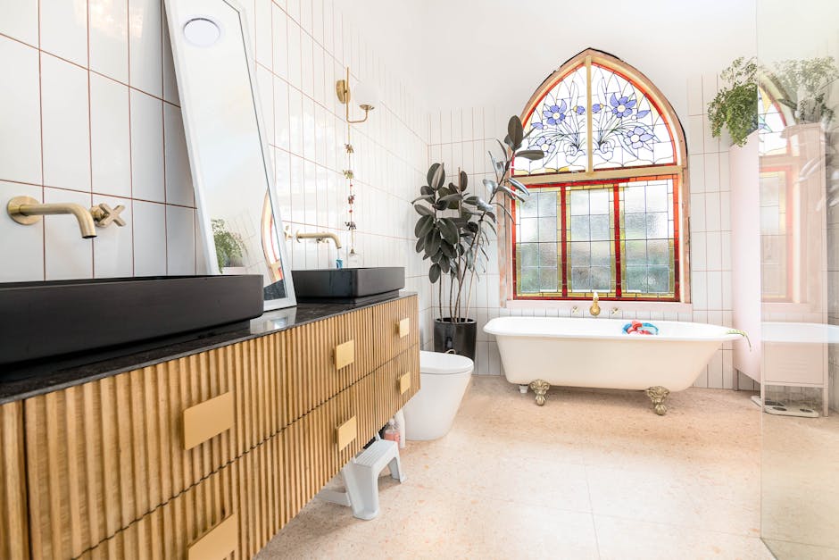 A spacious, bright bathroom featuring a vintage-style white bathtub with ornate clawfoot legs positioned in front of a large, arched stained glass window with blue floral patterns and red frames. To t