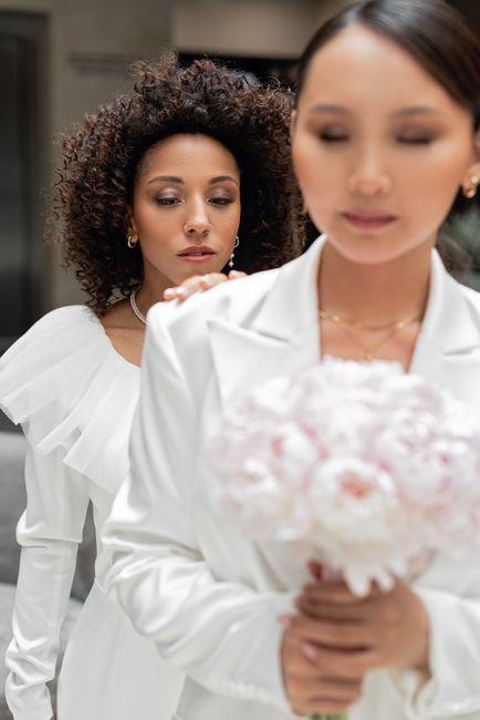 A woman with fair skin and dark hair, dressed in a white blazer, is holding a bouquet of soft pink and white flowers, possibly roses and peonies, wrapped in pink tissue and placed in a clear vase. She