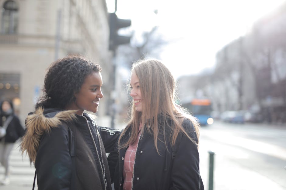 A candid street scene showing two women engaged in a friendly conversation on West Green Road N15. One woman, with curly dark hair, is wearing a black coat with a fur-lined hood, while the other, with