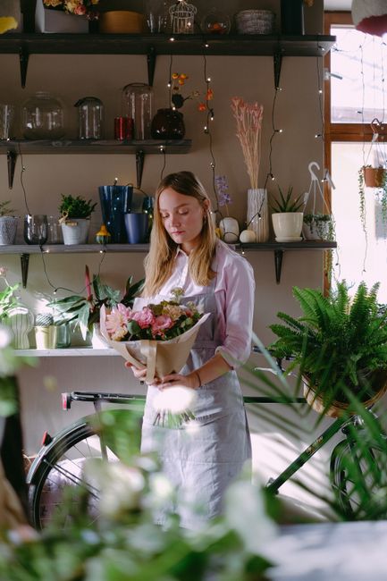 A young woman with long, light brown hair, wearing a white shirt and apron, gently holds a bouquet of fresh flowers wrapped in brown craft paper in a cozy indoor setting. The bouquet includes pink ros