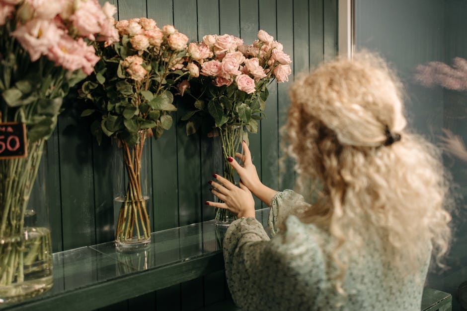 A woman with curly blonde hair, wearing a patterned top, arranging a bouquet of pale pink roses in a glass vase at a florist shop. The arrangement is placed against a dark green wooden wall, with seve