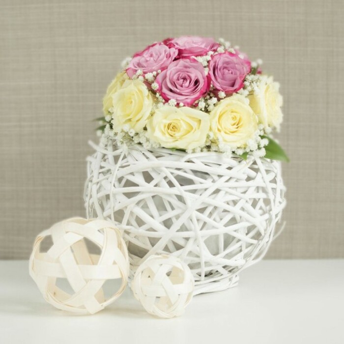 In this serene image, a delicately styled floral centerpiece evokes the feel of a small, elegant wedding reception tucked away in a cosy West Green venue. The eye is immediately drawn to the compact dome of roses, where around a dozen blooms are nestled closely together to form a lush, rounded silhouette. Half of the flowers are in a gentle blush to lavender-pink hue, their petals soft and velvety, some still slightly cupped while others open wider in graceful spirals. The remaining roses are a creamy blend of pale yellow and ivory, adding a buttery warmth that contrasts subtly yet harmoniously with the cooler pinks. Sprinkled throughout are diminutive white blossoms of baby's breath, their airy clusters filling tiny gaps between the roses and giving the design a light, cloud-like softness. This romantic bouquet is perched atop a spherical vase made from white-painted wicker or slender branches. The vase's open-weave pattern twists and crosses in an intricate lattice, creating a sculptural, almost globe-like form that feels modern yet rustic, just the sort of piece you might imagine on a chic table near Chestnuts Park. The vase sits on a spotless white tabletop, its brightness matching the clarity of the vessel. To the left are two decorative rattan-style balls in natural, pale wood tones, one larger, one smaller, which mirror the woven motif and add a sense of balance and rhythm to the display. The backdrop is a calm, neutral beige wall with a soft, fabric-like texture, keeping the focus on the floral artistry. Gentle, even lighting eases across the scene, enhancing the pastel colours and fine textures, making this arrangement an ideal inspiration for wedding table styling, anniversary dinners, or refined home décor throughout West Green.