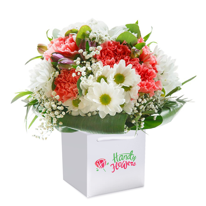 Presented by Florist West Green, this opulent bouquet stands tall in a minimalist white square gift bag, perfectly suited for same-day gifting across the Woodlands Park and West Green Road neighbourhoods. The arrangement brims with soft coral-pink and peach carnations, recognized for their luxuriously ruffled petals and comforting presence. White daisies, each dotted with a vivid lime-green center, playfully intersperse among the carnations, injecting a lively contrast. Throughout the composition, baby's breath drapes with lightness, its tiny ivory blossoms fluttering like confetti, while rich, emerald foliage and glossy broad leaves provide an elegant base, curving gently around the flowers. Tucked subtly within the bouquet are several closed buds, suffused with a hint of purple and promising to open soon, enhancing depth and dimension. The white bag's clean construction, complete with sturdy white ribbon handles, makes for a practical yet stylish means of delivery throughout West Green and neighbouring communities. The purity of the white background clarifies every colour and detail, highlighting the florist's artistry and the freshness of each stem. Ideal as a thoughtful gift for celebrations like birthdays, anniversaries or heartfelt congratulations, this bouquet encapsulates the joy and professional touch unique to West Green's local floral scene.