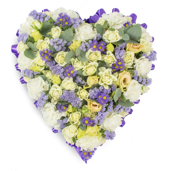 This meticulously arranged heart-shaped floral tribute is presented on a crisp white background, its form and coloration intended to offer solace and quiet beauty. The edge is defined by a continuous, rich purple satin ribbon, folded and placed to create a smooth, formal outline that accentuates the dense floral interior. Within that border, cream and pale yellow roses dominate-some tightly furled, others opening to reveal layers of silky petals with faint green tinges near the bud scales-offering a luxurious, tactile centre. Generous clusters of ruffled white carnations are threaded through the roses, supplying a soft, traditional texture that bathes the arrangement in calm. Punctuating the lighter hues are sprays of lavender statice and small purple aster-like blooms with bright yellow centres, which provide concentrated notes of colour and a slightly wild, meadow-like contrast to the formal heart. Blue-green eucalyptus leaves are artfully placed to break up the floral mass, their rounded silhouettes and cool tones adding structure, a modern sensibility, and an implied subtle fragrance. Gentle, even studio lighting reveals the interplay of surface and shadow, emphasizing craftsmanship and the peaceful mood intended for funeral services, wakes, or graveside remembrances. For residents of West Green and nearby Tottenham Green, the tribute carries a local sensibility-quiet, unassuming, and respectful-serving as a dignified way to honour a loved one with composure and heartfelt sentiment.