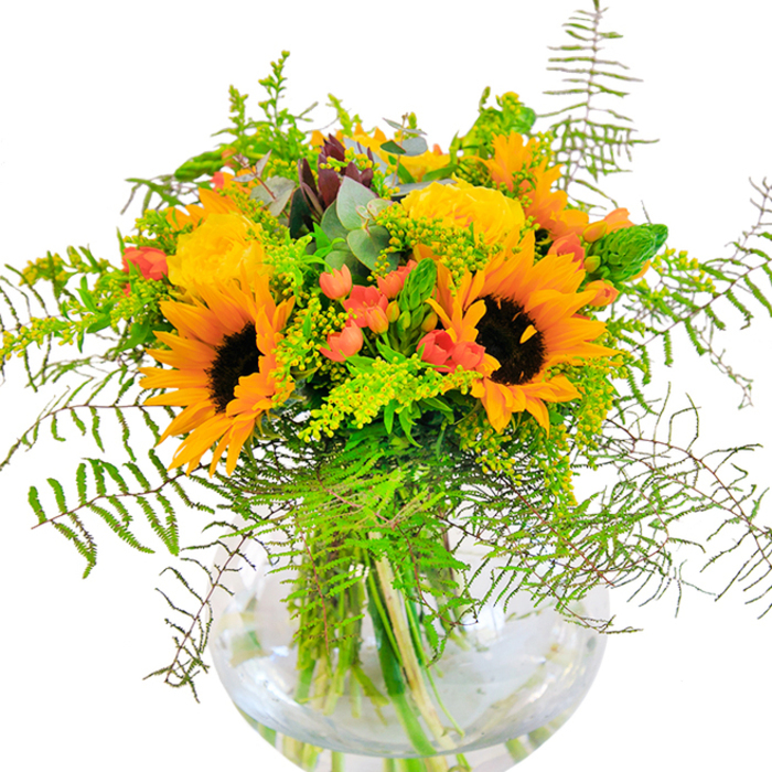 Overflowing with radiant beauty and the promise of sunny days in West Green, this floral arrangement captivates from every angle. Sunflowers, their vibrant yellow petals and rich brown centers unfurling in joyful profusion, set the tone at the heart of the bouquet-each bloom a miniature sun, evoking the open skies over nearby Downhills Park. Interwoven among them, buttery yellow garden roses open in lush spirals, their fragrance delicate yet uplifting, and their velvety curls a tribute to the area's historic gardens. Clusters of ruby-red Hypericum berries nestle playfully within the foliage, glossy and bright, offering a lively counterpoint to the dominant golden hues. The surrounding greenery-fine-textured fern fronds, slender spikes of solidago, and dusky eucalyptus leaves-spills outward in harmonious motion, giving the bouquet a sense of natural abundance and wild grace. The clear, rounded glass vase cradles every stem, its transparency displaying the bouquet's inner architecture and the palpable freshness that defines every arrangement crafted for West Green's homes. As light streams over the ensemble, shadows mingle softly with color, conjuring the essence of summer celebrations, birthdays, or welcoming gatherings. This bouquet is not just a floral centerpiece but a heartfelt expression of happiness-a piece of West Green delivered with passion and artistry.